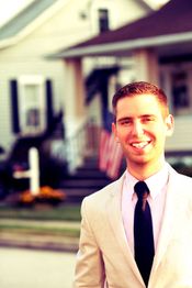 Smiling man in a suit standing outside a house with an American flag in the background.