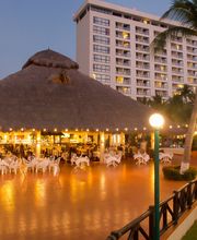 Terraza iluminada con palapa y sillas blancas junto a un edificio alto y palmeras al atardecer.