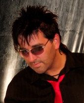 Mann mit Sonnenbrille, schwarzem Hemd und roter Krawatte vor grauem Hintergrund.
