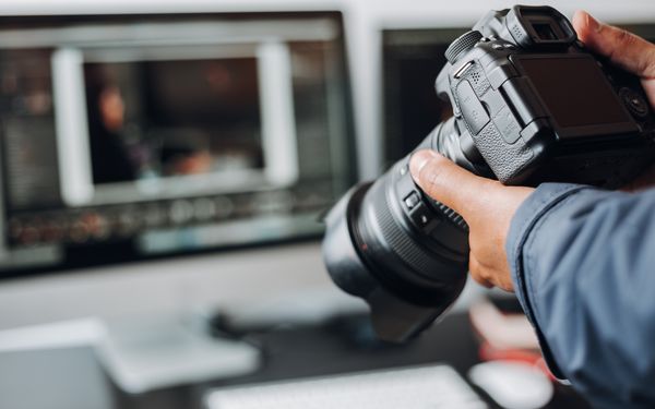 Fotograf hält einen Kamera in der Hand, im Hintergrund ist ein Bildschirm mit einer geöffneten Bildbearbeitungsprogramm zu sehen.