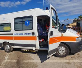 Ambulanza parcheggiata con porta aperta su una strada. Cielo sereno e alcune bandiere sullo sfondo.