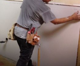 Worker installing drywall with tools in a belt indoors.