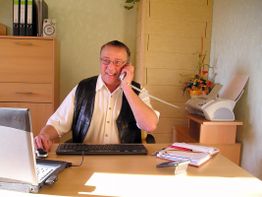 Mann telefoniert lächelnd im Büro, arbeitet am Laptop, Drucker und Ordner im Hintergrund.