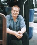 Ein Mann sitzt vor einem blauen Fahrzeug, lächelnd und in einem blauen Hemd gekleidet.