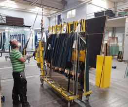 Person in Fabrik bedient Maschine, die große Glasscheiben trägt. Schutzbekleidung ist sichtbar.