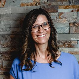 Frau mit Brille und lockigem Haar lächelt vor einer Steinwand in blauem Oberteil.