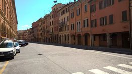 Strada cittadina con auto parcheggiate e case colorate sullo sfondo, cielo sereno.