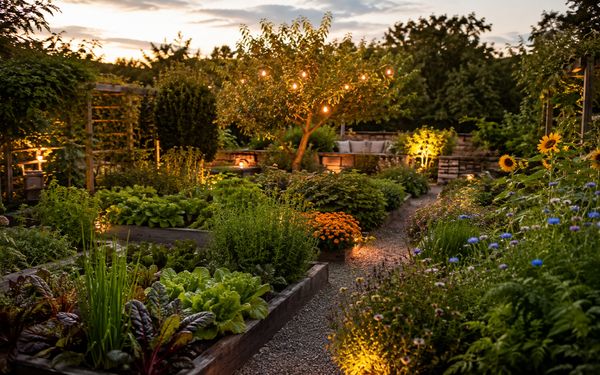 Moderner Bauerngarten mit Gemüse- und Kräuterbeeten, bunten Blumen, Obstbaum und stimmungsvoller Gartenbeleuchtung im warmen Abendlicht.