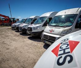 Mehrere weiße Transporter mit "FUCHS"-Logo parken nebeneinander auf einem Schotterplatz.