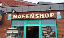 Hafen-Shop mit grüner Tür und Zeitungsständern davor, auf einem roten gemauerten Gebäude.