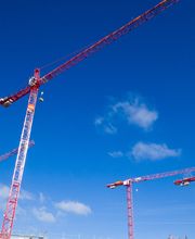 Drei rote Baukräne vor blauem Himmel mit wenigen Wolken.