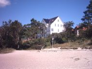 Weißes Haus hinter Bäumen am Sandstrand unter klarem blauem Himmel.