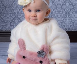 Ein Baby in weißem Outfit mit rosa Häschen, Schleifenstirnband, sitzt auf Holzboden.