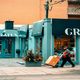 A person pushes a cart with boxes past blue storefronts on a street.