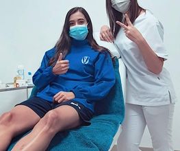 Dos personas con mascarillas, una sentada en una camilla y otra de pie haciendo señal de paz.