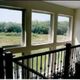 View through large windows overlooking green trees and fields from a staircase with a black railing.