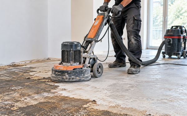 Ein Bodenleger entfernt mit einer Bodenschleifmaschine alte Klebereste vom Estrich, während ein angeschlossener Industriesauger den entstehenden Staub absaugt.
