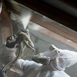 Worker in protective gear sprays insulation in building interior.