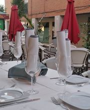 Mesa exterior de restaurante con manteles blancos y copas, rodeada de sillas y sombrillas rojas.