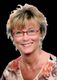 Eine lächelnde Frau mit Brille und Blumenbluse vor schwarzem Hintergrund.