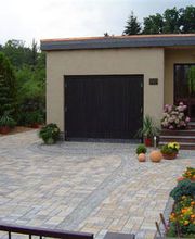 Einfahrt mit gepflastertem Boden, Garage, Topfpflanzen und Blumenbeeten vor einem Haus.