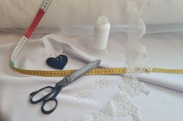 Nähzubehör mit Schere, Maßband, Garn, Spitze und Stecknadeln auf weißem Stoff.