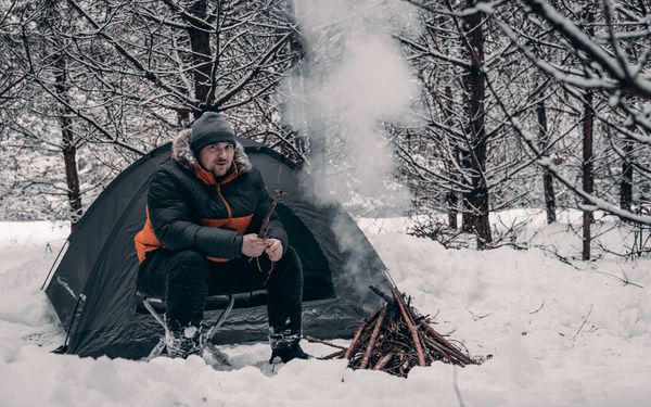 Ein Wanderer macht in einem verschneiten Wald neben einem blauen Zelt ein gemütliches Feuer.