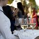 Server holding a tray with wine glasses at an outdoor event with people in the background.