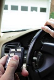 Hands holding a remote control and steering wheel in a car, aiming at a closed garage door.