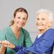 Caregiver in green scrubs smiling, holding hands with an elderly woman in a wheelchair.