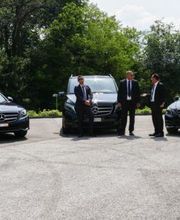 Drei schwarze Mercedes-Benz und zwei Männer in Anzügen auf einem Parkplatz.