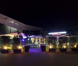 Nachtaufnahme eines beleuchteten Restaurants mit Bäumen in großen Pflanzkübeln davor.