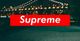 Red "Supreme" logo over a blurred night cityscape with a lit bridge in the background.