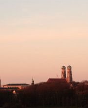Münchener Skyline im Abendlicht mit klar erkennbarem Kirchturm und blassen Himmel im Hintergrund.