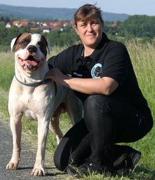 Frau kniet neben großem, weiß-braunem Hund auf einem Weg, ländliche Umgebung im Hintergrund.