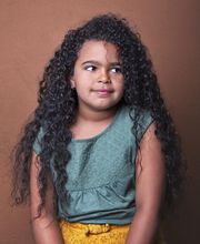 Niña de cabello rizado, con camiseta verde y pantalón amarillo, mirando hacia arriba.