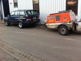 Blaues Auto mit Anhänger vor einer Halle geparkt, Anhänger enthält orangefarbenes Gerät.