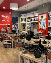 Tienda de utensilios de cocina con sartenes y electrodomésticos en estanterías de madera.