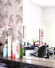 Friseursalon mit Spiegeln, Haarsprays, Bürsten und Blumen auf einer Kommode vor einer gemusterten Wand.