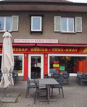 Zweistöckiges Gebäude mit Pizzeria und Kebab-Shop, Tische und Stühle draußen, Reklameschild rechts.