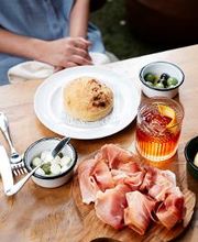 Mesa com pão, azeitonas, bebida e tábua de frios; pessoa ao fundo, sentada.