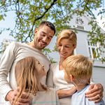 Happy family smiling in front of a house, surrounded by green leaves.