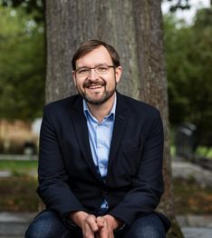Ein lächelnder Mann mit Brille sitzt vor einem Baum, trägt ein Sakko und ein blaues Hemd.