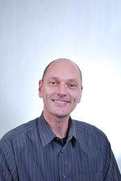 Ein lächelnder Mann in einem gestreiften Hemd vor einem hellen Hintergrund.