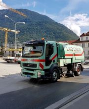 Straßenkehrmaschine fährt auf Straße vor Baugelände und Bergen im Hintergrund.