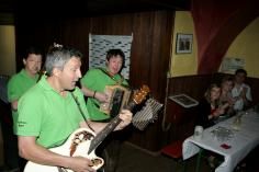 Gruppe in grünen Shirts spielt Gitarren und Akkordeon in einem Raum, während Leute am Tisch sitzen.