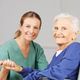 Caregiver in green scrubs smiling with an elderly woman in a wheelchair, both holding hands.