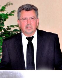 Ein Mann im Anzug steht vor einem Pult, mit einer Pflanze im Hintergrund.