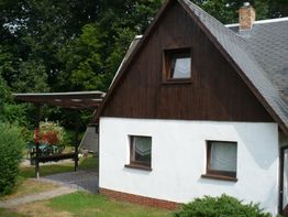 Kleines Haus mit Holzdach, weißen Wänden, umgeben von grüner Natur und Gartenmöbeln.