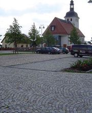 Kopfsteinpflaster-Parkplatz mit parkenden Autos, Bäumen und einer Kirche im Hintergrund.